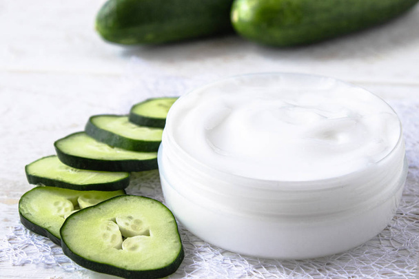 Face cream in a white jar with slices of fresh cucumbers on a white wooden background. - Fotografie, Obrázek