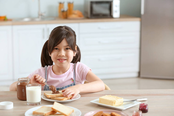 Забавная маленькая девочка ест вкусные тосты с шоколадом, распространяющимся на кухне
 - Фото, изображение