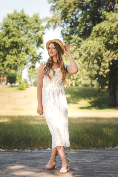 volledige lengte weergave van mooi meisje in witte jurk en stro hoed staande op weide en op zoek weg - Foto, afbeelding