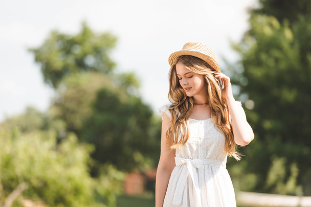beautiful girl in white dress touching straw hat and looking down - Photo, image