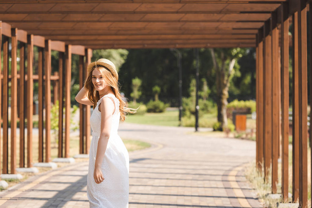 beautiful girl in white dress touching straw hat while walking near wooden construction and looking at camera - Photo, Image