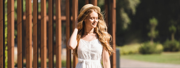 panoramic shot of beautiful girl in white dress touching straw hat and smiling with closed eyes  - Foto, Imagen