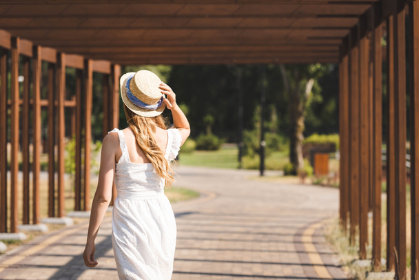 vista trasera de la chica en vestido blanco tocando sombrero de paja mientras camina por el camino cerca de la construcción de madera
  - Foto, Imagen