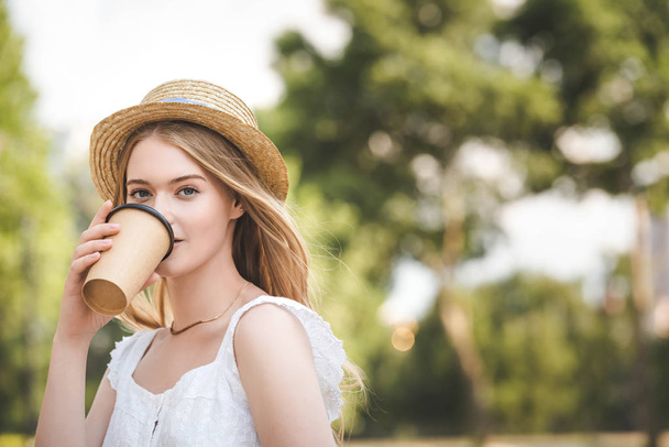 beautiful girl in white dress and straw hat holding paper coffee cup and looking at camera - Photo, Image