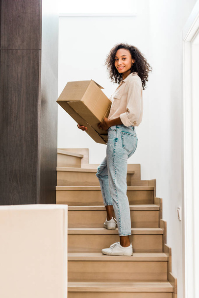 full length view of african american woman walking upstairs while holding box and looking at camera  - Photo, Image