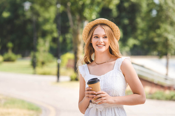 beautiful girl in white dress and straw hat holding paper cup, smiling and looking away - Фото, зображення