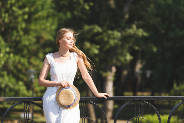 beautiful girl in white dress holding straw hat while leaning on handrail and standing with closed eyes - Zdjęcie, obraz