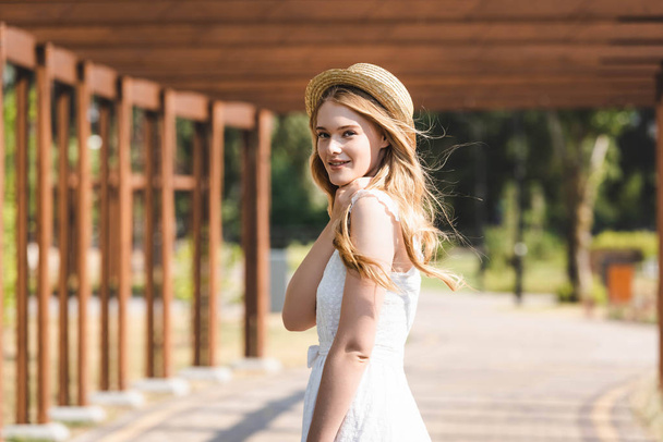 selectieve focus van mooi meisje in witte jurk en stro hoed wandelen in de buurt van houten constructie en kijken naar camera - Foto, afbeelding