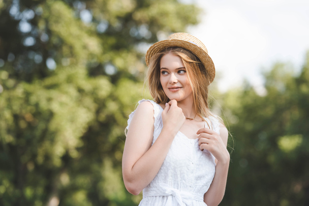επιλεκτική εστίαση του όμορφου κοριτσιού σε ψάθινο καπέλο και λευκό φόρεμα κοιτάζοντας μακριά - Φωτογραφία, εικόνα