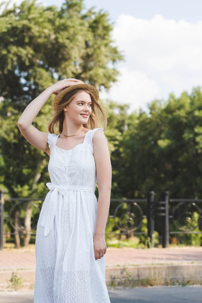 hermosa joven en vestido blanco tocando sombrero de paja mientras sonríe y mirando hacia otro lado
 - Foto, imagen