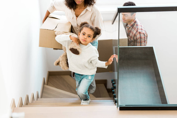 abgeschnittene Ansicht einer afrikanisch-amerikanischen Frau, die mit einer Schachtel nach oben geht, während die Tochter Spielzeug hält und der Vater der Mutter hinterherläuft  - Foto, Bild