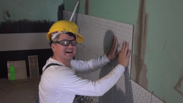 Facharbeiter mit gelbem Helm legt Fliesen an Wand und schaut in Kamera - Filmmaterial, Video