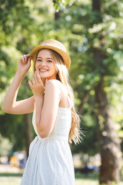 selective focus of beautiful young woman in white dress and straw hat smiling and looking at camera - Фото, изображение