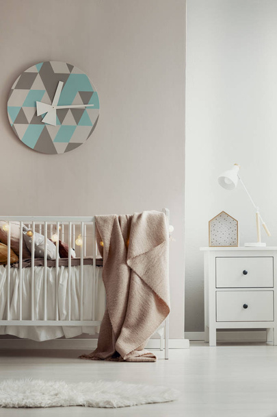 Modern, geometric clock hanging on beige wall in warm baby room interior with white crib. Real photo - Foto, Imagem