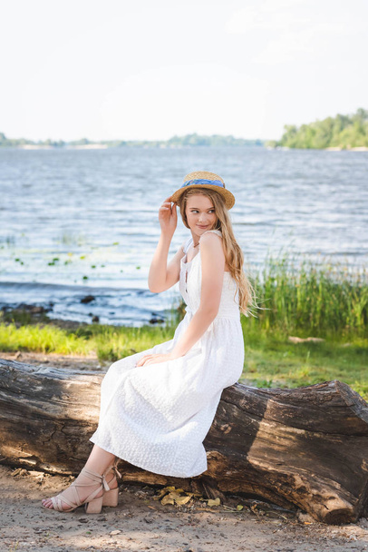 beautiful girl in white dress and straw hat sitting on trunk of tree on river shore and looking away - Photo, Image