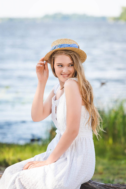 beautiful girl in white dress touching straw hat while sitting on trunk of tree on river shore and looking away - Photo, Image