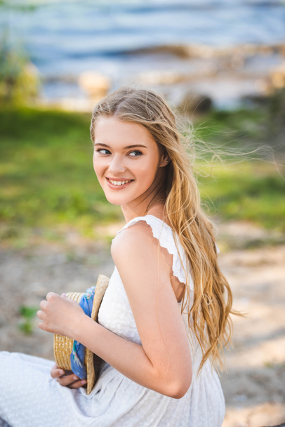portrait shot of beautiful girl in white dress holding straw hat, smiling and looking away - Foto, Imagem