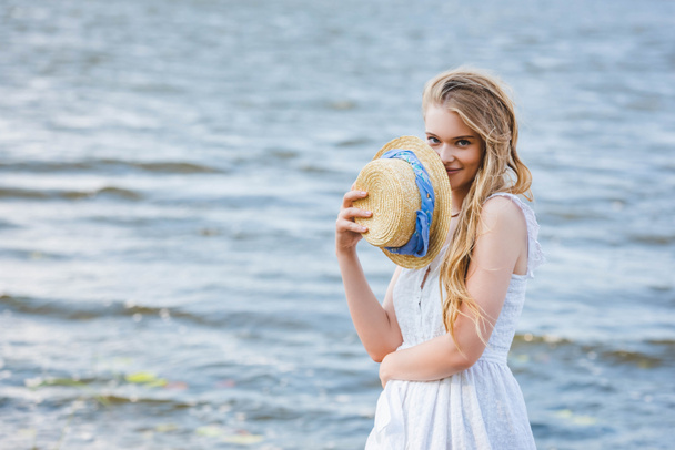 beautiful girl holding straw hat near face, smiling and looking at camera - Foto, Imagem