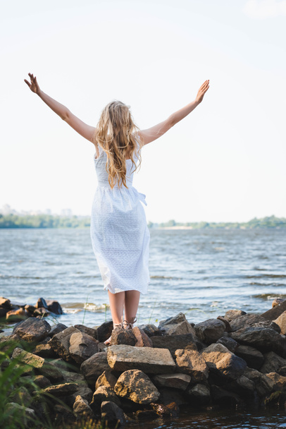 back view of girl in white dress standing on rocky river shore with hands in air - Valokuva, kuva