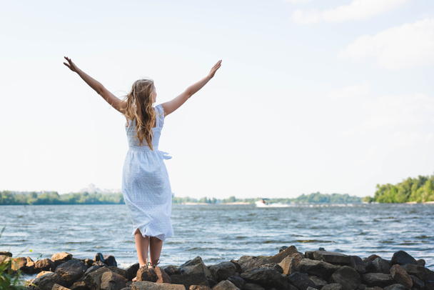 full length view of girl in white dress standing on rocky river shore with hands in air - Photo, Image