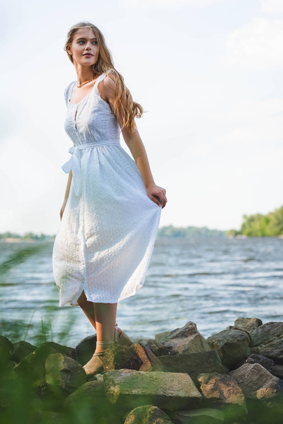 foco seletivo da menina bonita em vestido branco andando na costa do rio rochoso e olhando para longe
 - Foto, Imagem