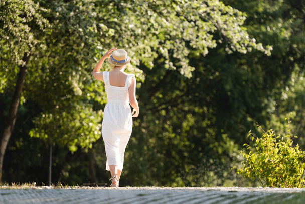back view of girl in white dress touching straw hat while walking in park - Fotografie, Obrázek
