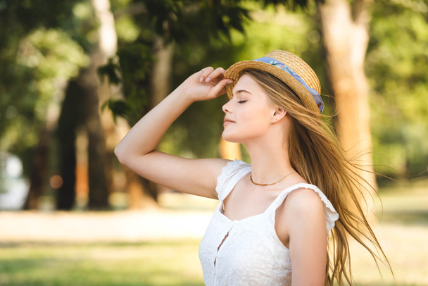 beautiful young girl in white dress touching straw hat while standing in park with closed eyes - 写真・画像