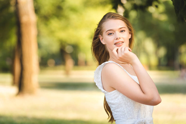 vista frontal de la hermosa joven en vestido blanco posando mientras está de pie en el parque y mirando a la cámara
 - Foto, imagen