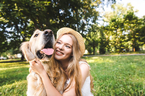 beautiful girl in white dress and straw hat hugging golden retriever while sitting on meadow, smiling and looking at dog - Photo, Image
