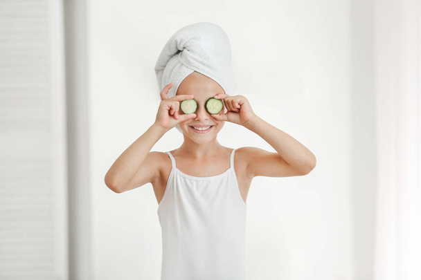 Cute little girl covering eyes with cucumber slices in bathroom - Foto, Imagen