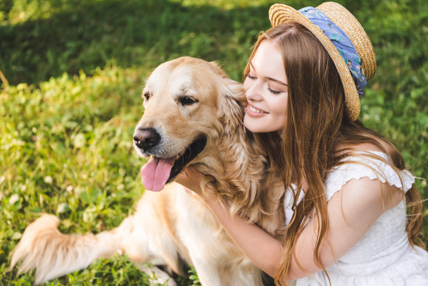 beautiful girl in white dress and straw hat hugging golden retriever while sitting on meadow and looking at dog - Photo, image