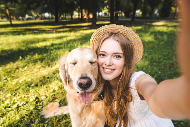 νεαρό κορίτσι με λευκό φόρεμα και ψάθινο καπέλο αγκαλιάζει Γκόλντεν Ριτρίβερ και παίρνοντας selfie ενώ κάθονται στο λιβάδι, χαμογελώντας και κοιτάζοντας την κάμερα - Φωτογραφία, εικόνα