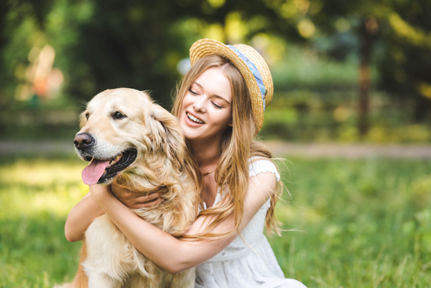selective focus of beautiful girl in white dress and straw hat hugging golden retriever and smiling while sitting on meadow - Φωτογραφία, εικόνα