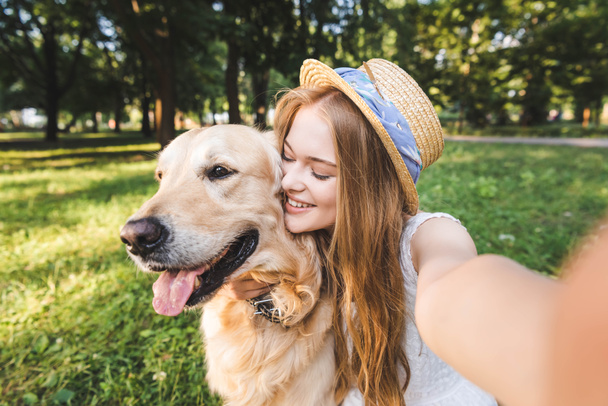 young girl in white dress and straw hat hugging golden retriever and taking selfie while sitting on meadow and smiling with closed eyes - Фото, изображение