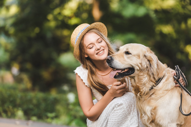  beautiful girl in white dress and straw sitting near golden retriever and smiling while looking at dog - Foto, imagen