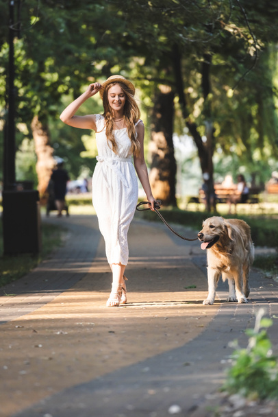 full length view of beautiful girl in white dress and straw hat smiling while walking with golden retriever on pathway and looking away - Foto, immagini