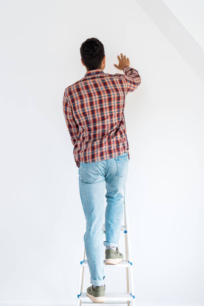 back view of man standing at ladder and looking at wall isolated on white - 写真・画像