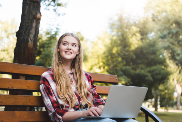 piękna dziewczyna w casual ubrania siedzi na drewnianej ławce w parku, za pomocą laptopa i patrząc z dala - Zdjęcie, obraz