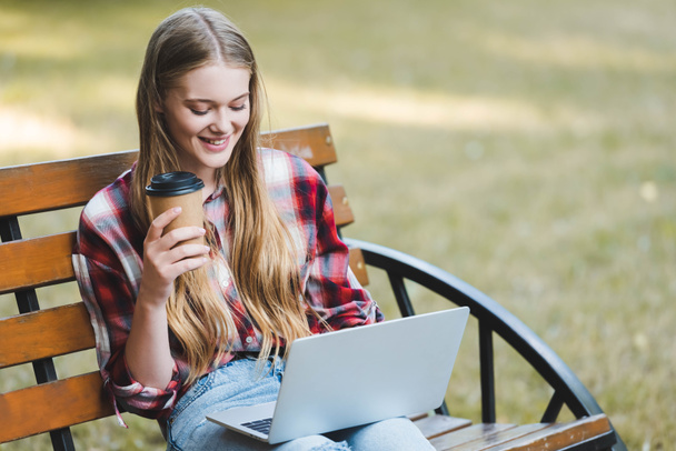 piękna dziewczyna w casual ubrania trzymając papier filiżankę kawy siedząc na drewnianej ławce w parku i za pomocą laptopa  - Zdjęcie, obraz