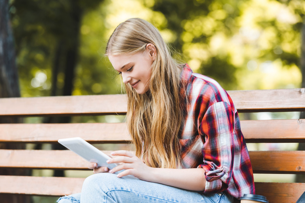 foyer sélectif de belle fille en vêtements décontractés assis sur un banc en bois dans le parc et en utilisant une tablette numérique
 - Photo, image