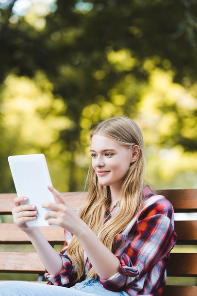 foyer sélectif de belle jeune fille en vêtements décontractés assis sur un banc en bois dans le parc et en utilisant une tablette numérique
 - Photo, image