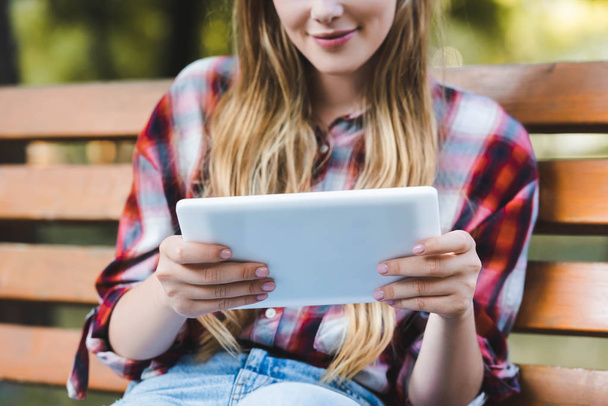 parkta ahşap bankta oturan ve dijital tablet kullanarak rahat giysiler genç kızın kırpılmış görünümü - Fotoğraf, Görsel