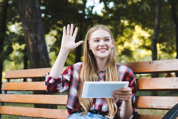 beautiful girl in casual clothes sitting on wooden bench in park, using digital tablet and waving hand - Foto, Imagem