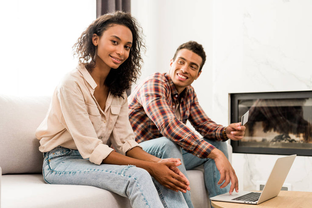 Africano casal americano sentado no sofá enquanto homem segurando cartão de crédito e mulher olhando para a câmera
  - Foto, Imagem