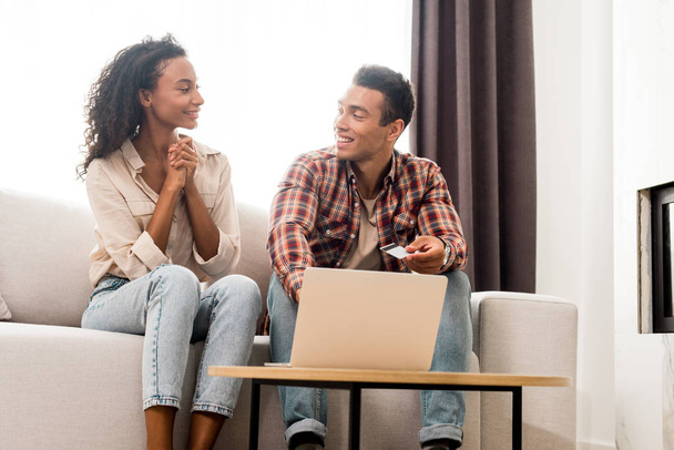 handsome man and african american woman looking at each other and sitting on sofa while husband holding credit card  - Zdjęcie, obraz