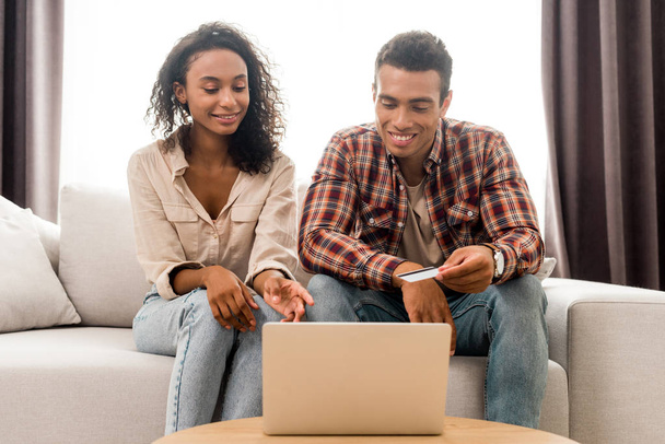 schöner afrikanisch-amerikanischer Mann und Frau schaut auf Laptop und sitzt auf Sofa, während Ehemann Kreditkarte hält  - Foto, Bild