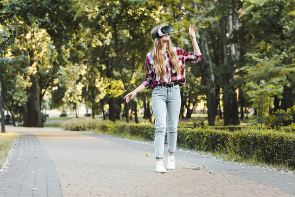 Вид в полный рост девушки в повседневной одежде с помощью гарнитуры VR во время прогулки в парке
  - Фото, изображение