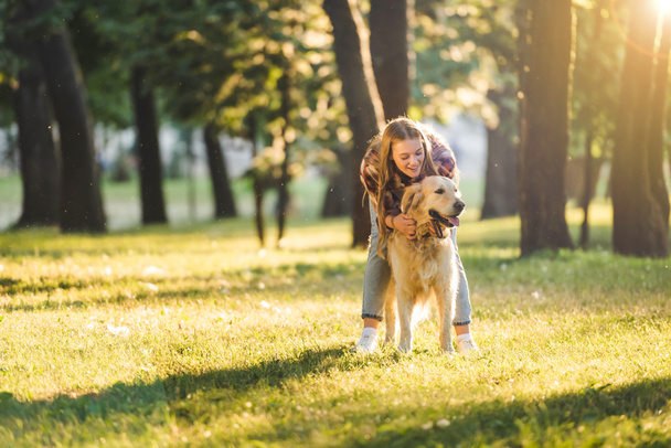 full length view of beautiful girl in casual clothes hugging golden retriever while standing on meadow in sunlight - Fotografie, Obrázek
