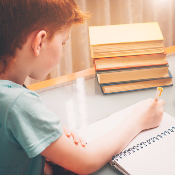 Red-haired boy writes in a notebook while doing school homework surrounded by books - Foto, Bild