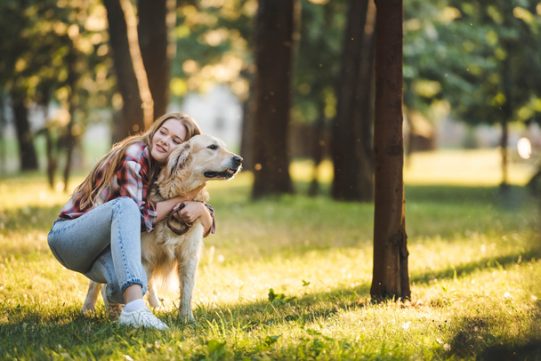 full length view of beautiful young girl in casual clothes hugging golden retriever while sitting on meadow in sunlight and looking away - Фото, изображение